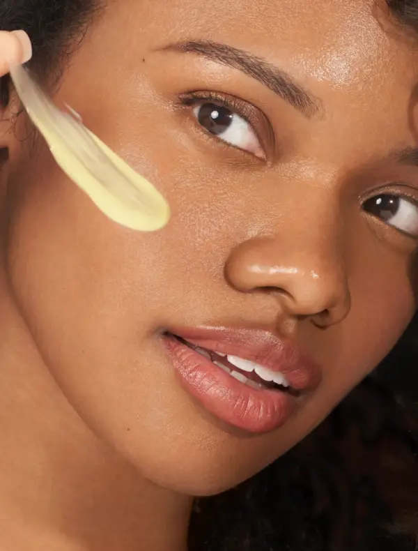 A close-up of a woman’s face with smooth skin as a hand applies Retinol+ 1.0 30ml cream to her cheek using a spatula.