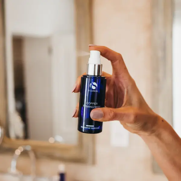 A hand holds the Copper Firming Mist 75ml, a blue spray bottle, highlighting this skin care essential in front of a blurred bathroom mirror and sink.
