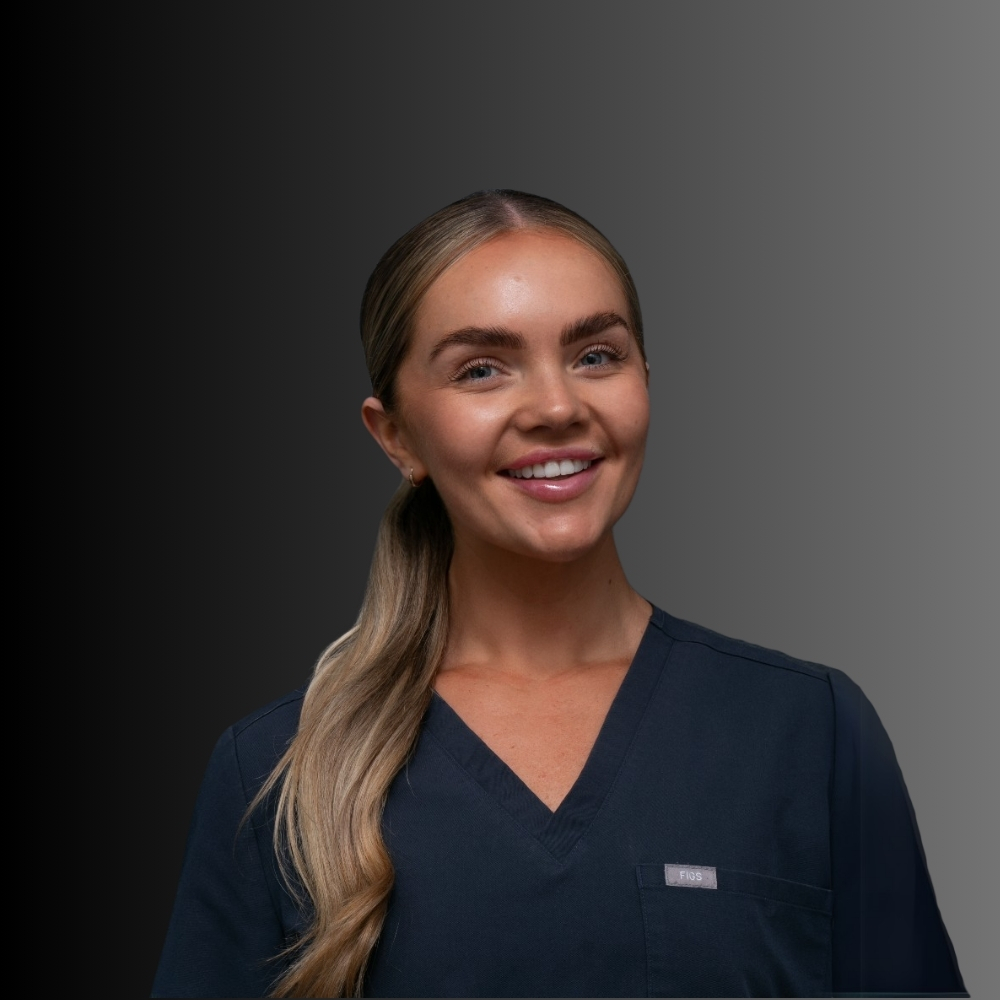 Zoe-2 - Kaya Cosmedica A woman wearing dark blue medical scrubs stands in front of a gradient black-to-gray background, smiling at the camera with her hair pulled back in a low ponytail.