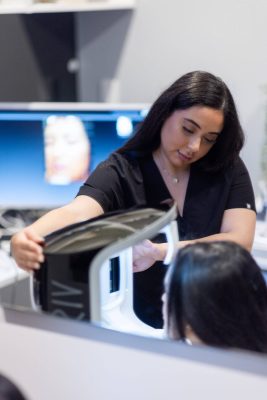 A woman with dark hair examines her reflection in a high-tech Visia mirror, adjusting equipment on a modern desktop, possibly in a beauty salon or medical clinic.